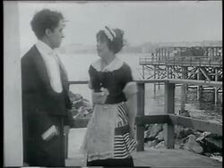 B/W 1915 man (Charlie Chaplin) + woman (Mabel Normand) on pier kicking each other in buttocks Stock Footage