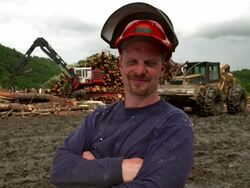 Medium shot. Portrait. Slight pan to the left as logger crosses his arms and smiles into the camera with logging site in background. Stock Footage