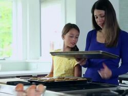MS DS daughter watching mom put tray of cookies in oven in kitchen of contemporary home Stock Footage