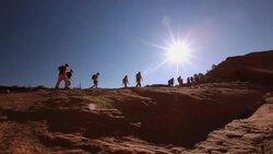 Wide shot of hikers hiking up steep red-rock hill in desert with blue skies and sun in Utah (pan) Stock Footage