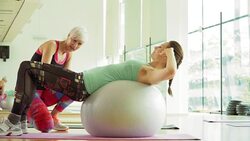 Personal trainer guiding woman doing sit-ups on fitness ball in gym Stock Footage