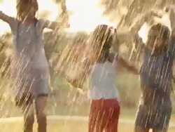 Three girls jumping in front of a lawn sprinkler in a park Stock Footage