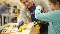 Hispanic father or woodworking workshop employee teaches elementary age girl how to use a power drill at woodworking camp for children Stock Footage