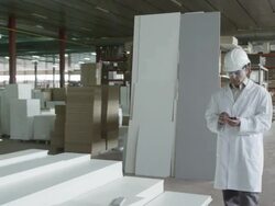 MS TS man in lab coat and hard hat walking along multi-story warehouse in furniture production, taking notes on handheld electronic device Stock Footage