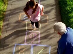 High angle wide shot senior woman waving as young girl plays Hopscotch on sidewalk Stock Footage