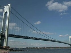 Small waves crashing on a beach boats sailing under the verrazano-narrows bridge in the background on a partly cloudy day Stock Footage