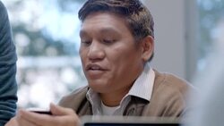 Older businessman shares smart-phone, confers with young group leader in office conference-room meeting Stock Footage