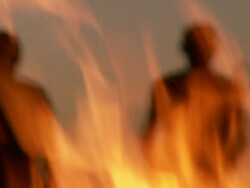 Out of focus silhouetted medium long shot of two bushmen walking towards camp-fire (shot through heat of flames) Stock Footage