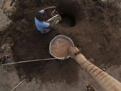 High Angle crane push-in - A camel on a rope drinks muddy water from a basin as a young man adds more water from a well. / Djibouti Stock Footage