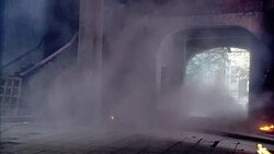 MEDIUM ANGLE OF COMBAT IN A RUNDOWN OR ABANDONED BUILDING. SEVERAL GUNMEN OR REBELS FIRE MACHINE GUNS FROM BEHIND A PILLAR IN ROOM AND FROM OUTSIDE BUILDING THROUGH LARGE OPEN DOORWAY. SMALL FIRES BURN ON FLOOR AND AN EXPLOSION FILLS THE ROOM WITH SMOKE. Stock Footage