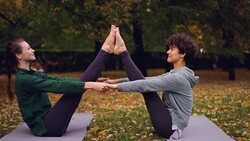 Attractive girls are doing yoga exercises in pair holding hands and putting feet together raising legs sitting on mat in park. Enjoyable training and people concept. Stock Footage