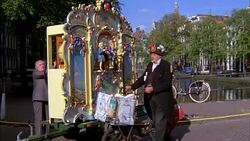 WIDE ANGLE OF MAN, ORGAN GRINDER WITH PUPPET SHOW ON SIDEWALK NEAR CANAL. STREET PERFORMER. Stock Footage