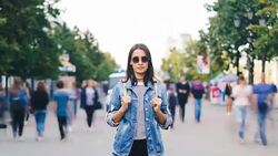 Time-lapse portrait of attractive brunette in sunglasses standing in the street by herself and looking at camera when men and women are passing by on autumn day. Stock Footage