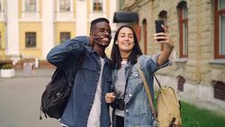 Happy tourists friends with backpacks are taking selfie using smartphone showing hand gestures v-sign and thumbs-up standing together in the street in beautiful city. Stock Footage