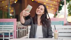 Beautiful girl with long dark hair is taking selfie and posing for smartphone camera sitting in street cafe alone. Modern lifestyle, youth and gadgets concept. Stock Footage