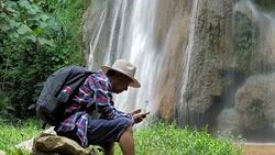 Senior man Asian traveler sitting enjoying and selfie with smart phone in front of the waterfall. Lifestyle Travel, Technology and Relaxation concept Stock Footage