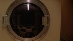 MEDIUM ANGLE ESTABLISH OF YELLOW WASHING MACHINE OR DRYER WITH CLEAR GLASS DOOR WITH METAL RIM. SEE REFLECTION OF BLACK AND WHITE CHECKERED FLOOR ON DOOR. SEE TALL RECTANGULAR OBJECTS IN REFLECTION. Stock Footage