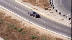 AERIAL TRACKING SHOT OF 1970'S BROWN TWO DOOR CAR DOWN FREEWAY OR HIGHWAY, PASSING OTHER CARS AS IT DRIVES OFF RAMP AND ONTO DIFFERENT FREEWAY. RURAL AREA. Stock Footage