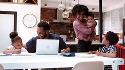 Father helps children with homework whilst mother holds baby daughter in kitchen at home Stock Footage