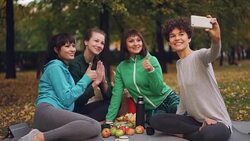 Attractive young ladies yogini are taking selfie using smartphone during picnic in park in autumn. Girls are posing and smiling having fun sitting on mats. Stock Footage