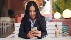 Smiling Asian girl student is resting in outdoor cafe and using smart phone touching screen and watching. Social media, modern technology and city concept. Stock Footage