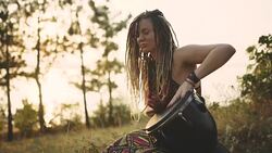 Beautiful young hippie woman with dreadlocks playing on djembe. Funky woman drumming in nature on an ethnic drum at sunset or sunrise Stock Footage