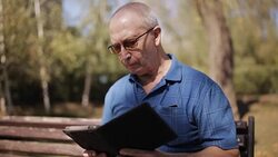 An elderly man with glasses reading an e-book on a Park bench Stock Footage