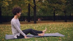 Slim young lady is doing sequence of yoga asanas sitting on mat in park alone on autumn day with beautiful grass and trees around her. Recreation and sport concept. Stock Footage