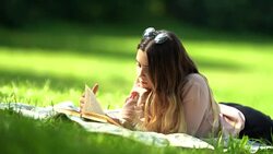 A Beautiful girl with long hair reads a book lying on the grass in the park on a sunny day Stock Footage