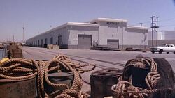 MEDIUM ANGLE OF DOCK OR HARBOR OR PIER. WOODEN POSTS WITH ROPES TIED IN NAUTICAL KNOTS ON THEM IN FOREGROUND. WAREHOUSE IN BACKGROUND. 1970'S ERA WHITE SEDAN ENTERS FRAME FROM RIGHT EDGE AND DRIVES OFF DOCK INTO OCEAN. CAR CRASH. CAR SINKS BELOW SURFACE. Stock Footage