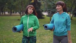 Cheerful young women are walking on grass and talking holding yoga mats after practice in city park. Communication, healthy lifestyle and friendship concept. Stock Footage