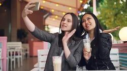Attractive girl is taking selfie with her Asian friend sitting in open-air cafe with drinks holding smartphone and posing with glasses. Friendship, technology and leisure concept. Stock Footage
