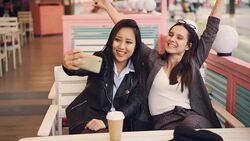 Carefree girls are taking selfie with take out drinks sitting in cafe together and using smartphone. Young women are posing, clinking glasses and showing hand gestures. Stock Footage