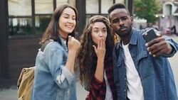 Good-looking young people Caucasian and African American tourists are taking selfie using smartphone posing with hand gestures having fun on summer day. Stock Footage