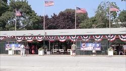 WIDE ANGLE OF ENTRANCE TO MONMOUTH PARK, HORSE RACING TRACK IN OCEANPORT, NEW JERSEY. SIGN FOR THE FOURTH OF JULY DERBY. PEOPLE, PEDESTRIANS, SPECTATORS. AMERICAN FLAGS. Stock Footage