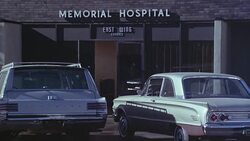 MEDIUM ANGLE ESTABLISH OF BRICK BUILDING HOSPITAL WING ENTRANCE WAY. SEE 'MEMORIAL HOSPITAL' AND 'EAST WING ENTRANCE' SIGNS OVER DOORS. SEE CHRYSLER STATION WAGON AND MERCURY COMET PARKED IN FRONT. SEE NURSE IN WHITE DRESS AND HAT EXIT FROM DOORS INTO PAR Stock Footage