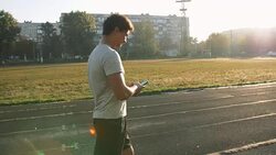 Young mixed race male sportsman track runner walking by race track with smartphone touching phone display before sports training at stadium Stock Footage