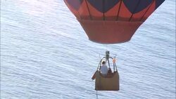 AERIAL TRACKING MEDIUM ANGLE OF HOT AIR BALLOON FLYING RIGHT TO LEFT OVER GLISTENING BLUE OCEAN. BALLOON IS MULTI-COLORED (YELLOW ORANGE AND RED). COULD BE LATE DAY OR EARLY MORNING. SEE MAN IN BASKET OF HOT AIR BALLOON. Stock Footage