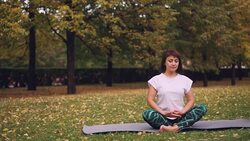 Pretty young woman is relaxing sitting in lotus pose on yoga mat in park and breathing resting after individual practice. Meditation and nature concept. Stock Footage