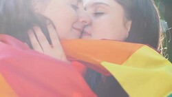 Two cheerful lesbians wrapped in rainbow flag hugging and kissing outdoors Stock Footage