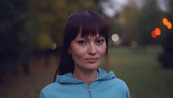 Portrait of beautiful young sportswoman with long dark hair and brown eyes looking at camera and smiling standing outdoors in autumn evening wearing hoody. Stock Footage