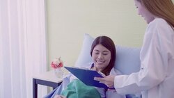 Beautiful smart Asian doctor and patient discussing and explaining something with clipboard in doctor hands while staying on Patient's bed at hospital. Medicine and health care concept. Stock Footage