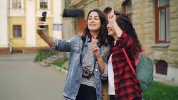 Cheerful girls foreign travelers are taking selfie using smartphone standing outdoors and posing with hand gestures showing v-sign and heart with fingers and laughing. Stock Footage